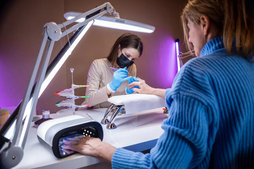 Manicurista profesional con guantes aplicando esmalte en uñas de cliente con lámpara LED y muestra de colores