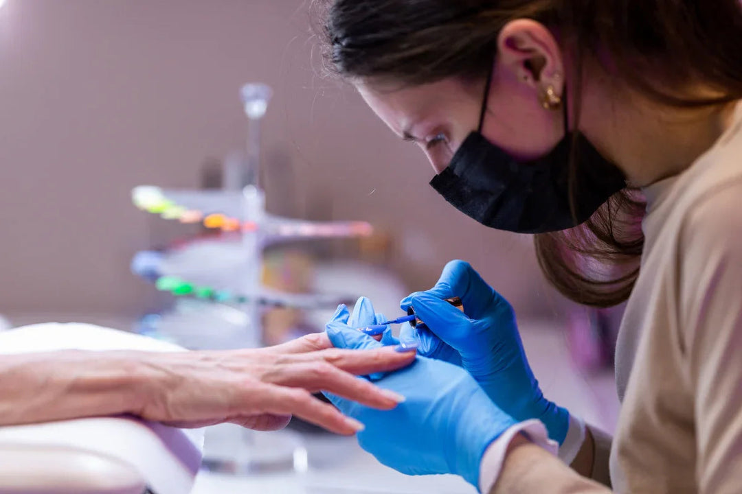 Manicurista con guantes azules y mascarilla aplicando esmalte azul en uñas de cliente en salón
