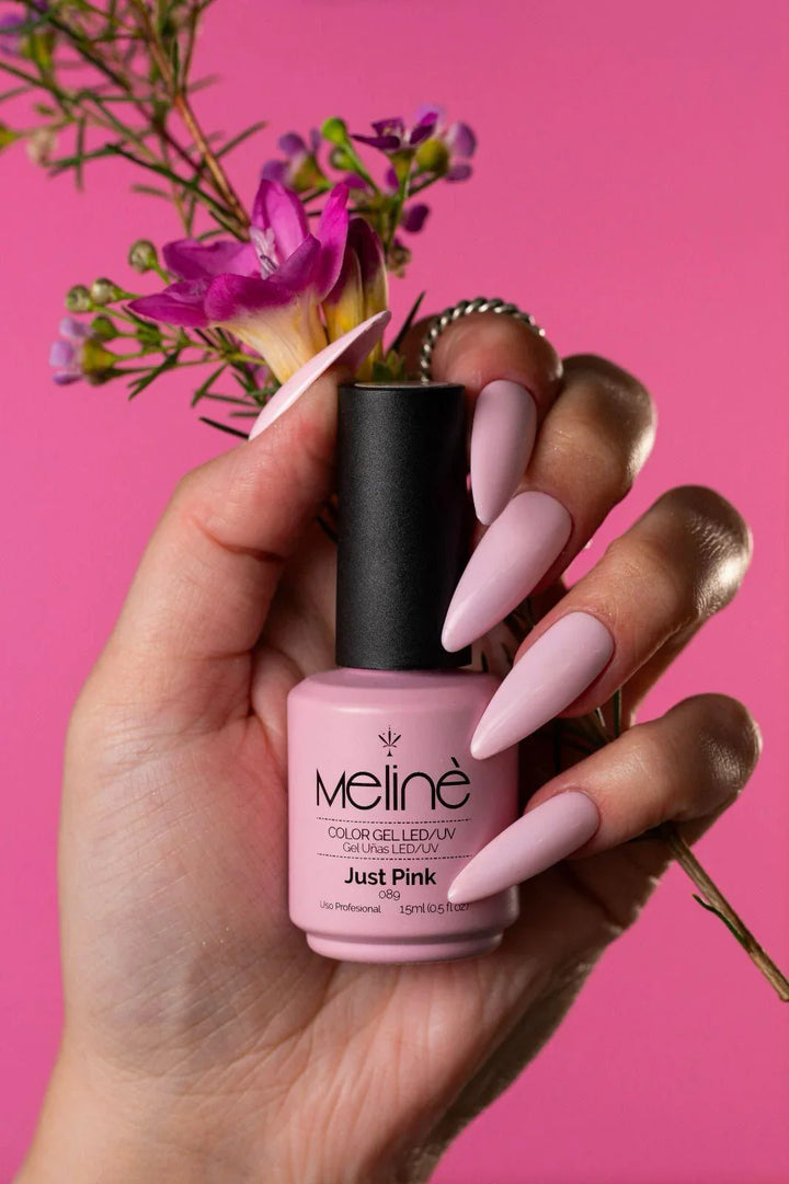 Mano con uñas largas en tono rosa sosteniendo esmalte semipermanente Melinè Just Pink, flores y fondo rosa