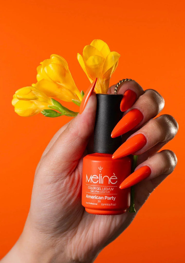 Mano con uñas largas naranja sosteniendo esmalte gel semipermanente Meline American Party y flores amarillas