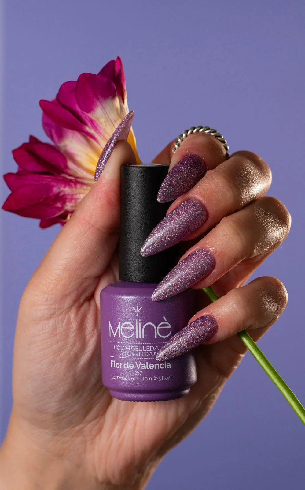 Mano con uñas largas en punta decoradas con esmalte purpura brillante sosteniendo botella Meline Flor de Valencia y flor morada
