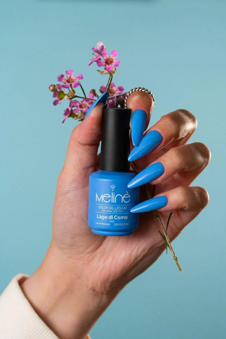 Mano femenina con uñas largas azul brillante sosteniendo esmalte semipermanente Meliné Lago di Como y flores moradas